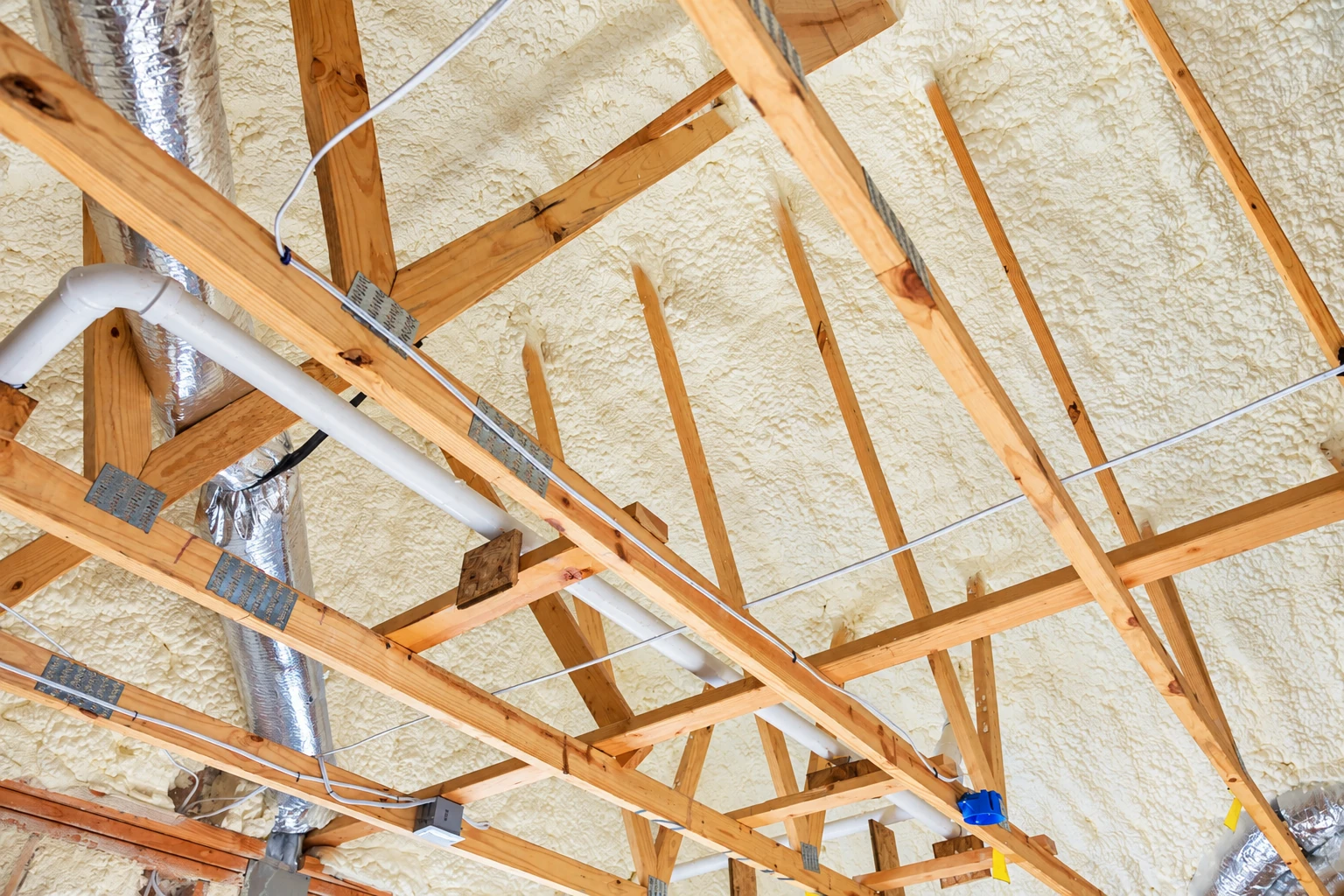 Attic rafters and roof deck coated in pale spray foam, with pipes and wiring crossing the frame.