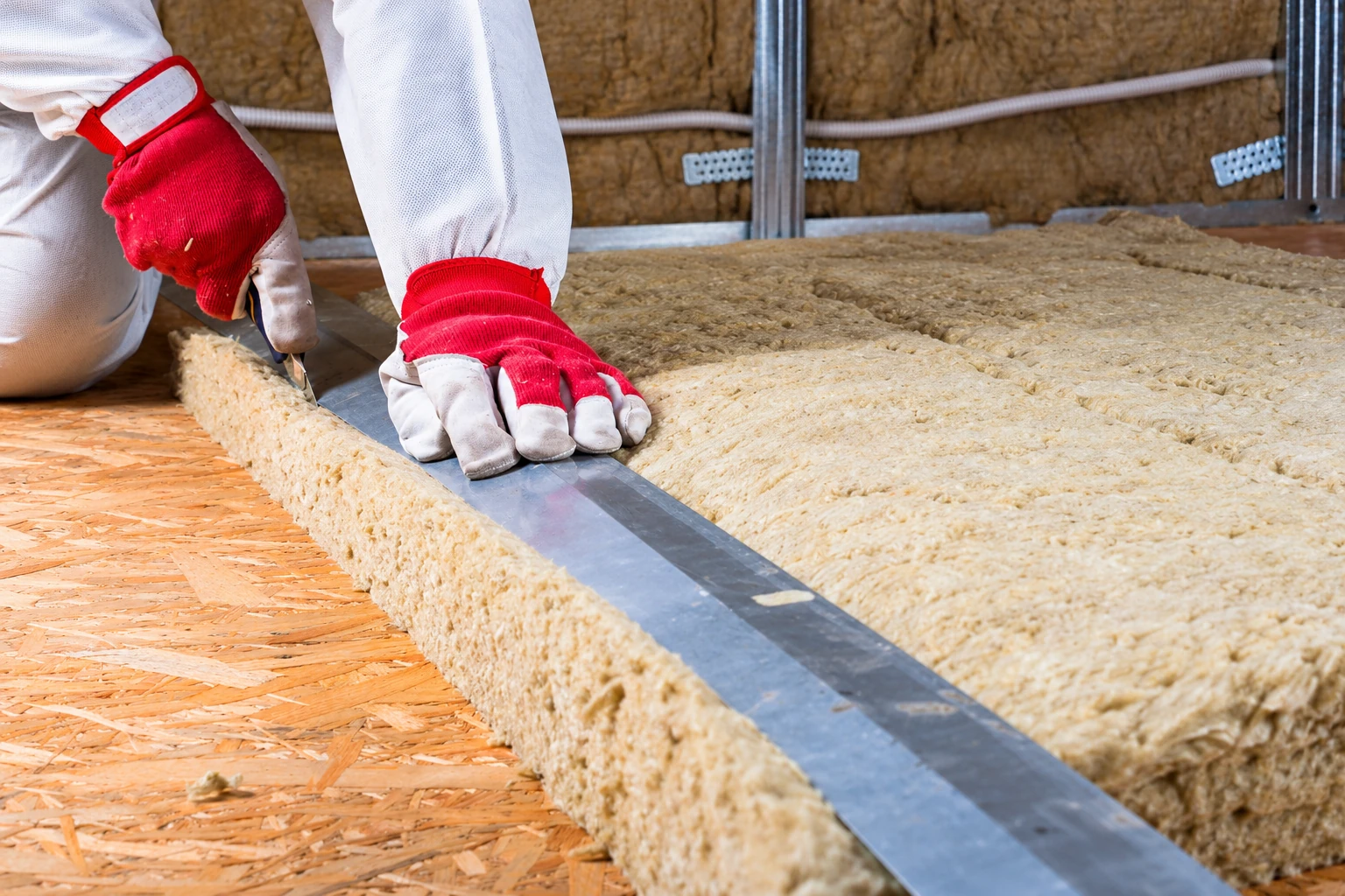 A worker in red gloves cuts a thick fiberglass insulation batt on an attic floor.