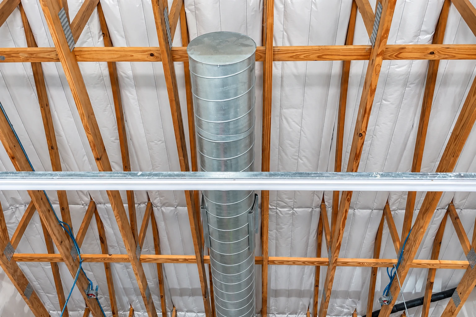Attic roof lined with white insulation between wooden rafters, with a large metal duct running down the center.