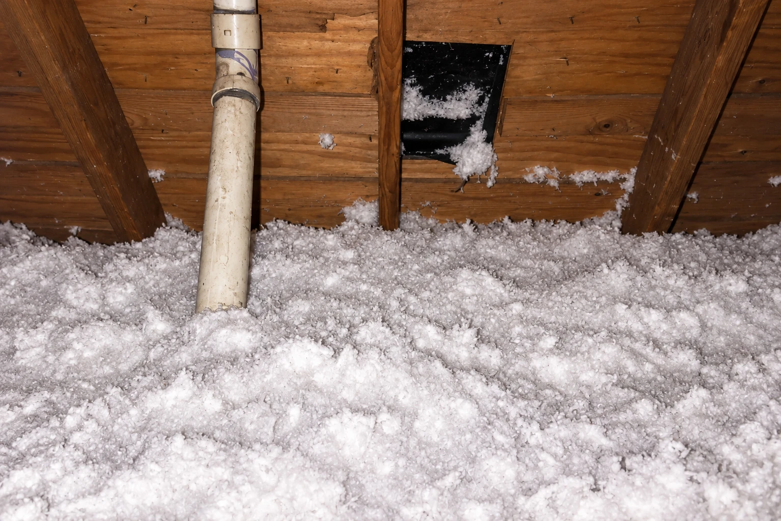 Attic floor covered in white blown-in insulation beneath exposed wooden rafters and a white vent pipe.