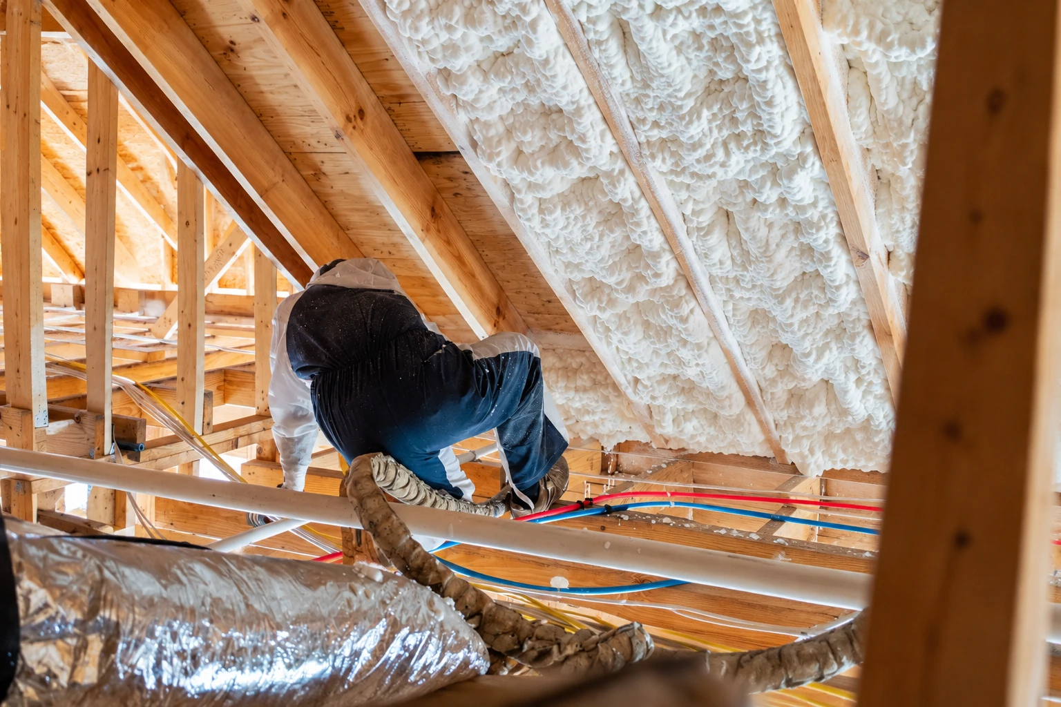 A worker crouches in a wooden attic, applying white spray foam insulation between roof rafters.